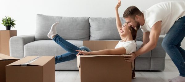 A woman is playfully sitting in a cardboard box and a man is pushing her in it across the loungeroom. They are both in blue jeans and white tees.