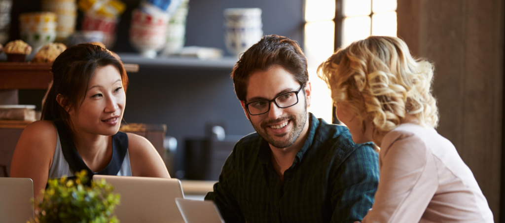 A couple are sitting at a table chatting with another woman. A laptop sits on the table.