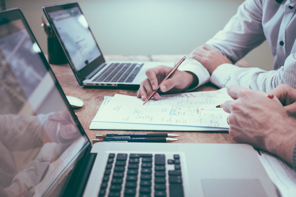 Two laptops sit on the desk and you can see two peoples hands which indicate they are working together over paperwork.